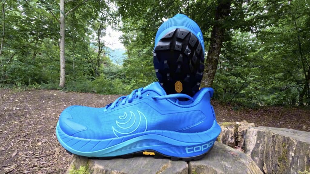 Bright blue Topo MTN Racer 4 trail running shoe on a tree stump, showing the Vibram Megagrip outsole and wide toe box in a forest setting.